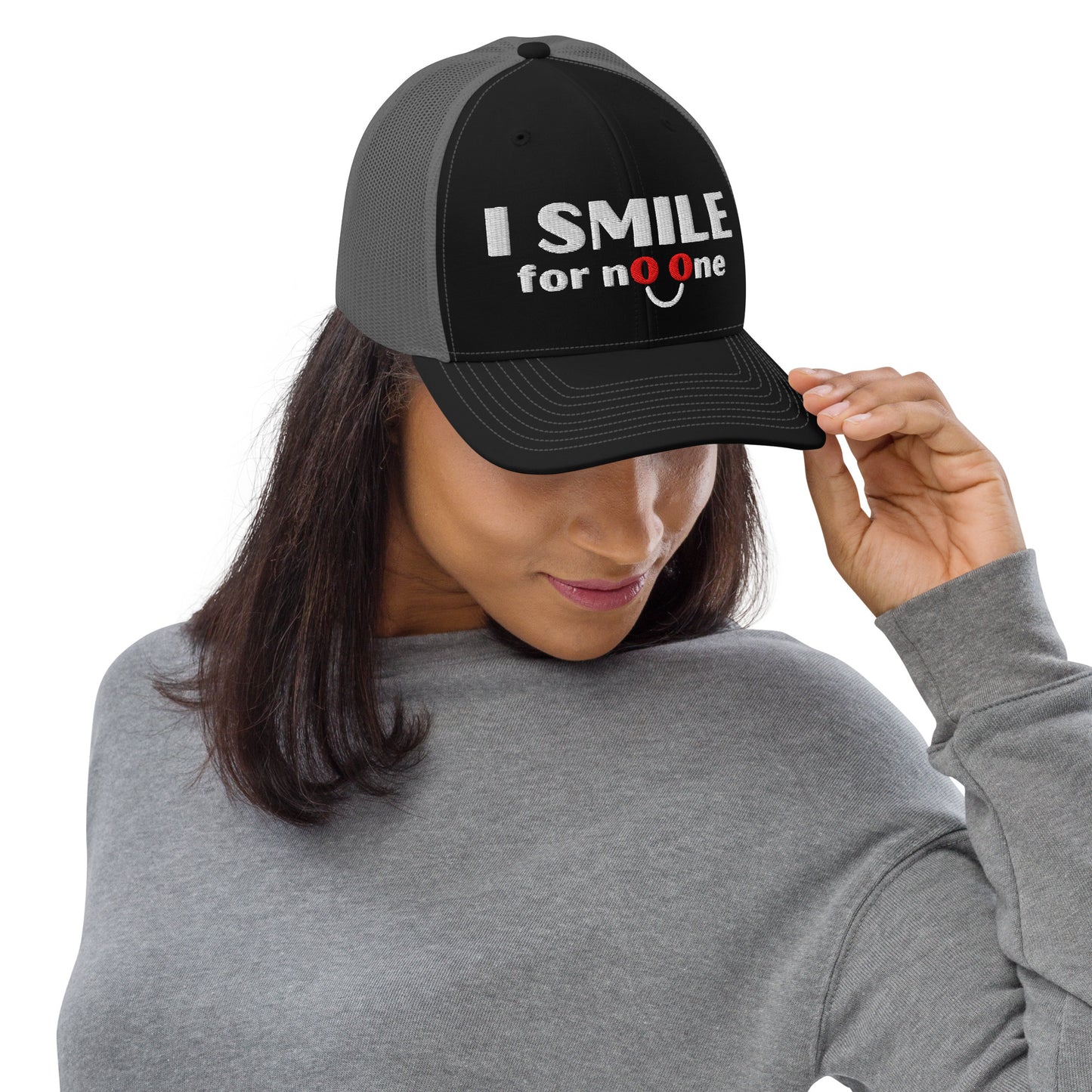 Woman in a black and grey trucker hat with embroidered text 'I Smile for No One' in bold white lettering. Structured cap with mesh back and adjustable snapback.