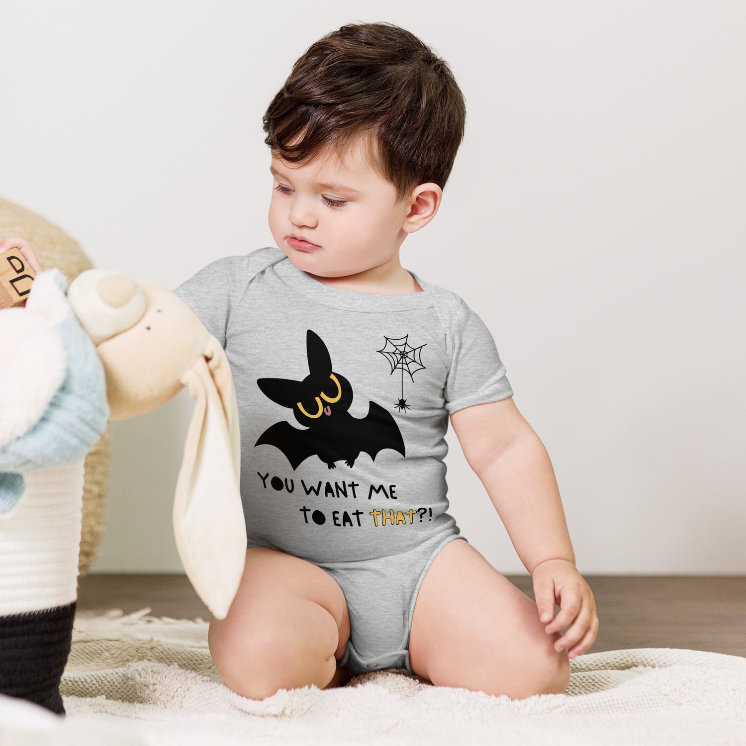 Toddler in a grey baby onesie with a cute bat next to a spider hanging off a spiderweb and caption "You Want Me To Eat THAT?!"